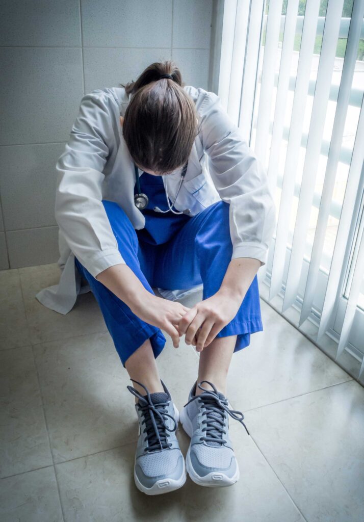 Woman in blue scrubs and white coat sits on floor of hospital with head down and arms on knees.