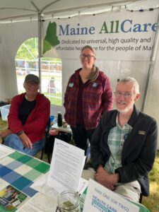 Two white men and one white woman sitting and standing behind a table inside a white tent, in front of a sign that says Maine AllCare, at the Common Ground Country Fair in Unity, Maine, September 2023.