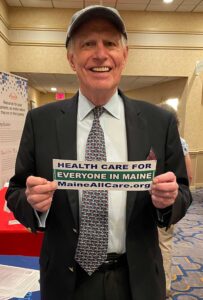 Close up of TR Reid, American filmmaker and author, holding a bumper sticker that says Healthcare for Everyone in Maine, Maine AllCare.org, at the Maine Medical Association annual meeting in September 2023.