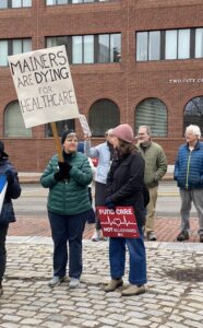Two women holding protest signs: Mainers are Dying for Healthcare, Fund Care, Not Billionaires