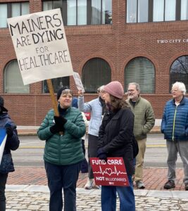 Two women holding protest signs: Mainers are Dying for Healthcare, Fund Care, Not Billionaires