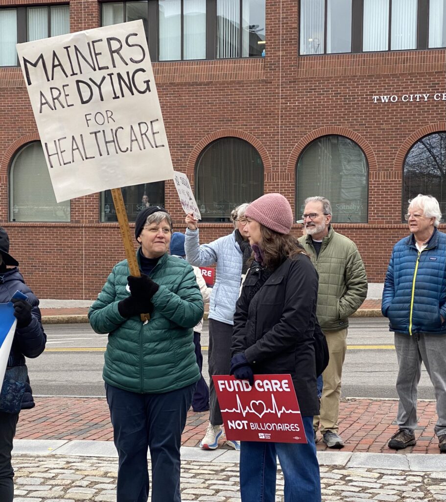 Two women holding protest signs: Mainers are Dying for Healthcare, Fund Care, Not Billionaires