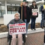 A white woman wearing winter wear, sitting in a wheelchair and holding a sign that reads "Love it! Improve it! medicare for All." Two others with healthcare focused signs stand behind her. They are in front of the federal building in Bangor, Maine.