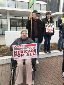 A white woman wearing winter wear, sitting in a wheelchair and holding a sign that reads "Love it! Improve it! medicare for All." Two others with healthcare focused signs stand behind her. They are in front of the federal building in Bangor, Maine.
